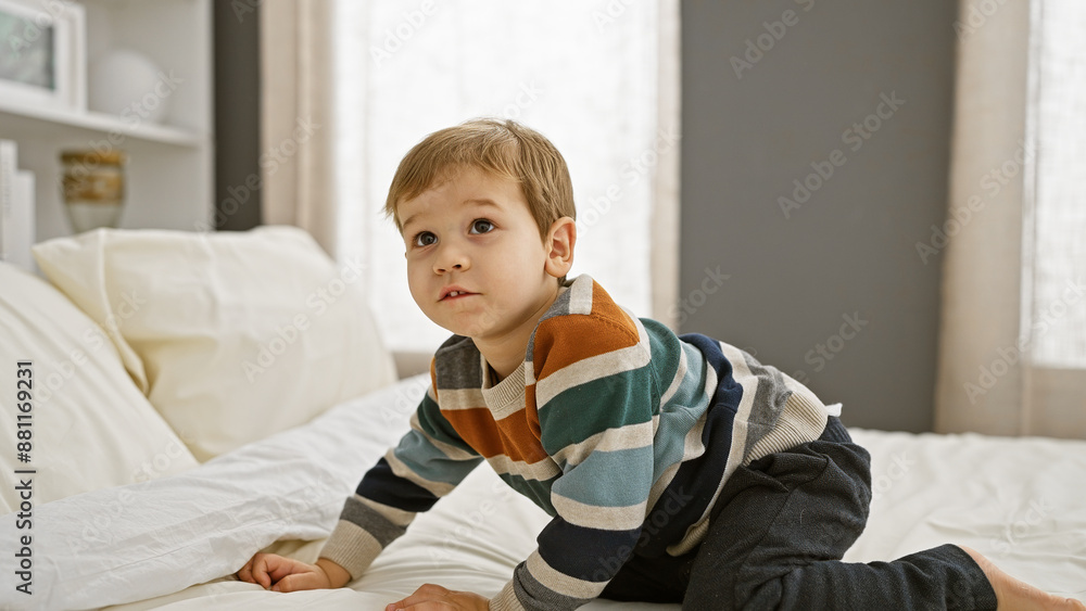 Blond toddler boy in striped shirt kneeling on white bed in a bright bedroom, looking curious. Blond toddler boy in striped shirt kneeling on white bed in a bright bedroom, looking curious.