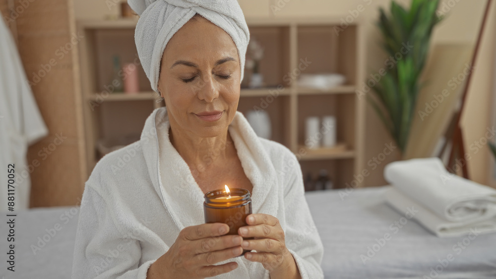 Middle-aged woman in a spa holding a lit candle, wearing a white bathrobe and towel on her head, relaxing in a serene wellness room with minimal decor and plants in the background Middle-aged woman in a spa holding a lit candle, wearing a white bathrobe and towel on her head, relaxing in a serene wellness room with minimal decor and plants in the background
