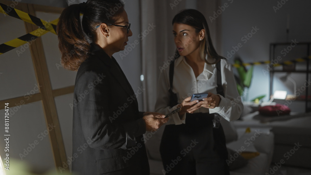 Fototapeta premium Two women investigating a crime scene indoors with one holding a phone and the other holding a notepad, surrounded by caution tape.