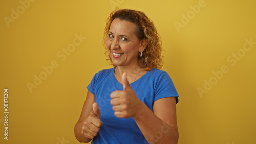 Elderly hispanic woman with curly hair and a blue shirt giving thumbs up while smiling, isolated over yellow background.
