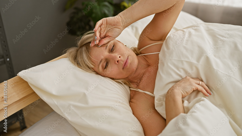 A blonde woman appears unwell resting in a bedroom, indicating sickness or headache in a home setting. A blonde woman appears unwell resting in a bedroom, indicating sickness or headache in a home setting.