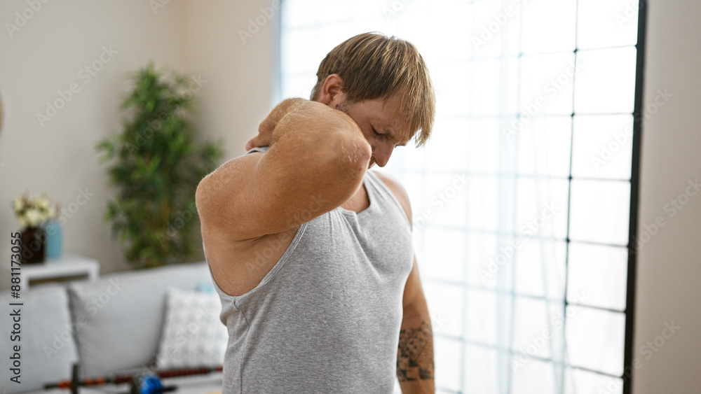 Obraz premium A young, caucasian man with a beard and blond hair in a tank top experiencing neck pain indoors at home.