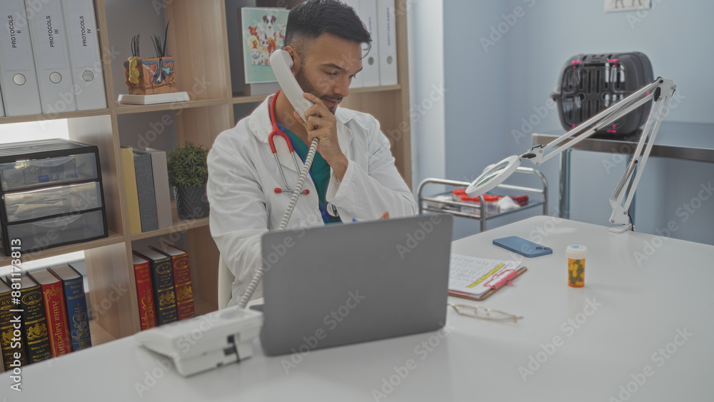 Obraz premium Young hispanic man in a veterinary clinic talking on the phone while working on a laptop in an indoor office setting with medical equipment and shelves in the background.
