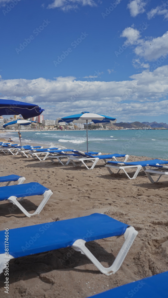 A serene beach scene with blue sunbeds, umbrellas, and waves against a backdrop of a cloudy sky and a cityscape. A serene beach scene with blue sunbeds, umbrellas, and waves against a backdrop of a cloudy sky and a cityscape.