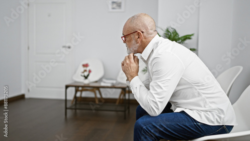 A pensive senior man with a beard and glasses sits alone in a minimalist white room, suggesting a quiet waiting area. photo