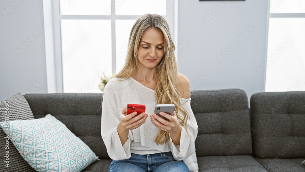 A young blonde woman compares smartphones while sitting comfortably in her modern living room. A young blonde woman compares smartphones while sitting comfortably in her modern living room.