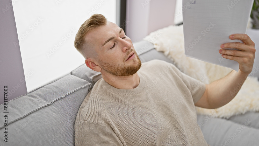 Obraz premium A caucasian man with a beard relaxes at home fanning himself with a document in a comfortable, modern interior.