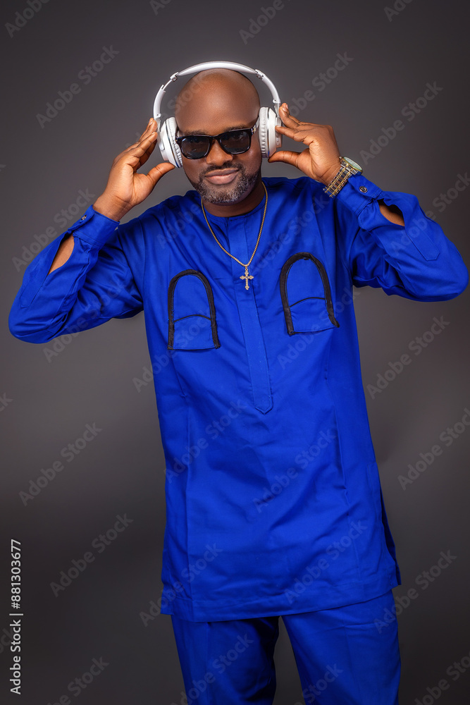 Close up shot of an African man in blue native attire wearing a gold chain and a wrist watch with hands on a headset on black shades smiling to the camera in a light dark background Close up shot of an African man in blue native attire wearing a gold chain and a wrist watch with hands on a headset on black shades smiling to the camera in a light dark background