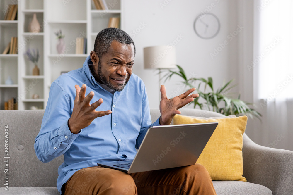 Man feeling frustrated while working on laptop at home. Sitting on couch with hands raised in exasperation. Home setting with bookshelf, plant, and clock in background. Man feeling frustrated while working on laptop at home. Sitting on couch with hands raised in exasperation. Home setting with bookshelf, plant, and clock in background.