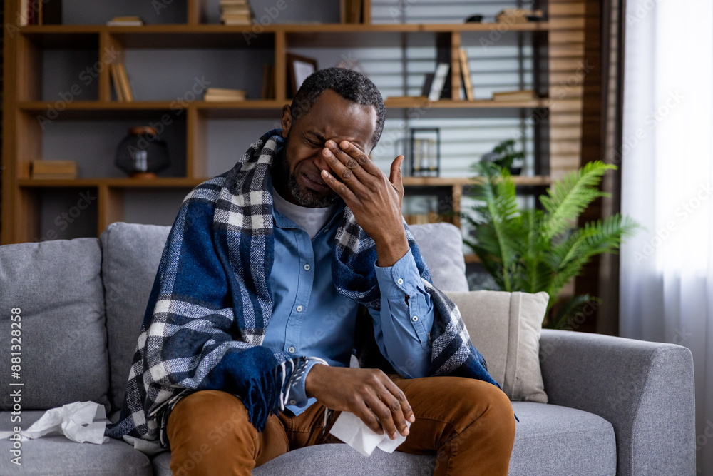 Man feeling unwell sitting on sofa covered with blanket holding tissues. Displaying symptoms of cold or flu, gripping face in discomfort. Home environment with bookshelves and plants in background Man feeling unwell sitting on sofa covered with blanket holding tissues. Displaying symptoms of cold or flu, gripping face in discomfort. Home environment with bookshelves and plants in background