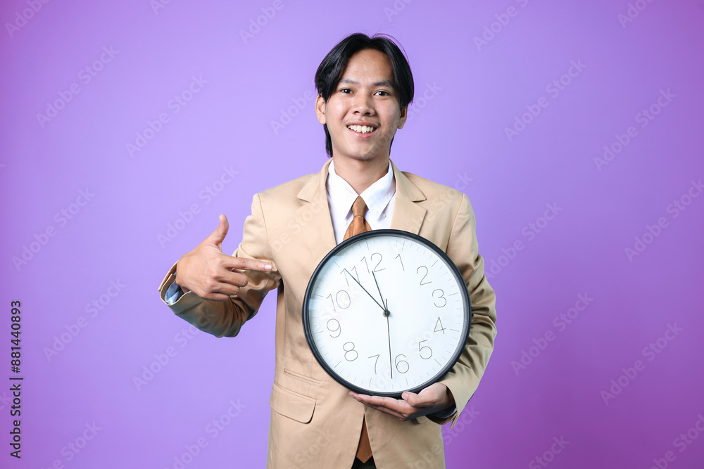 Friendly Young Asian Businessman Standing While Ponting And Holding Clock For Mockup Isolated On Purple Background Friendly Young Asian Businessman Standing While Ponting And Holding Clock For Mockup Isolated On Purple Background