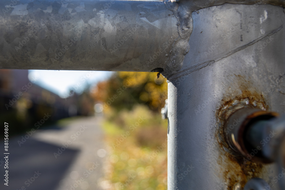 Close-up view captures rusty metal barrier with peeling paint - single water droplet poised at edge - against a backdrop of blurred autumnal trees lining a pathway. Taken in Toronto, Canada. Close-up view captures rusty metal barrier with peeling paint - single water droplet poised at edge - against a backdrop of blurred autumnal trees lining a pathway. Taken in Toronto, Canada.
