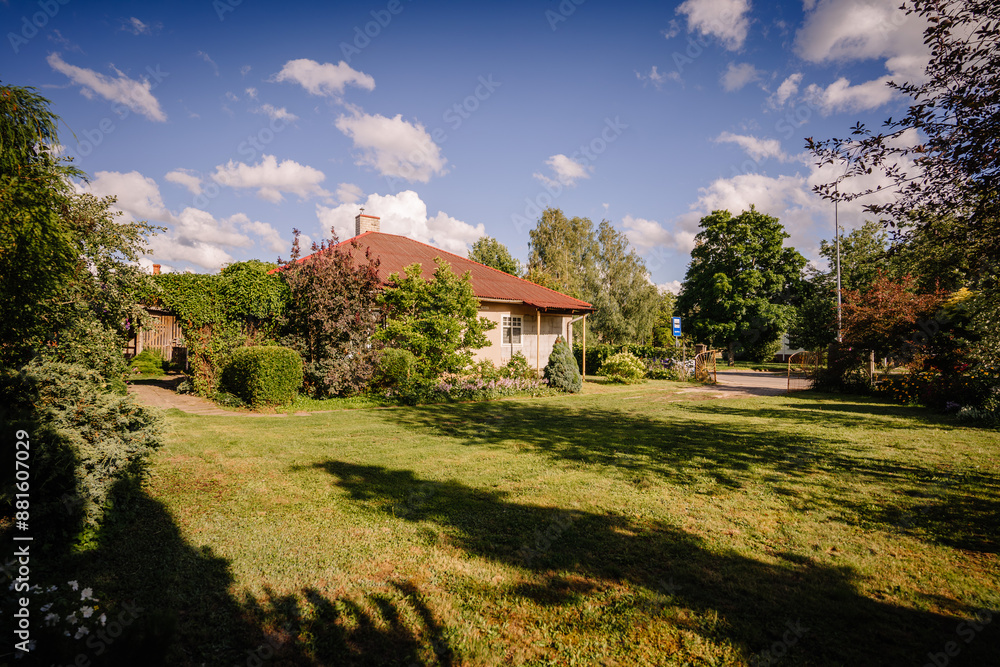 Valmiera, Latvia - July - 5, 2024 - A house with a red roof surrounded by a lush garden with various trees and shrubs, a sunny lawn, and a clear blue sky in the background. Valmiera, Latvia - July - 5, 2024 - A house with a red roof surrounded by a lush garden with various trees and shrubs, a sunny lawn, and a clear blue sky in the background.