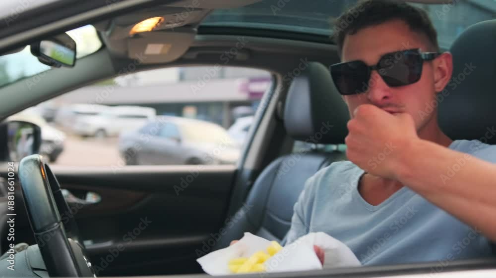 A young man sitting behind the wheel of a car eats French fries from a roadside fast food cafe on the road. Close up. Slow motion. A young man sitting behind the wheel of a car eats French fries from a roadside fast food cafe on the road. Close up. Slow motion.