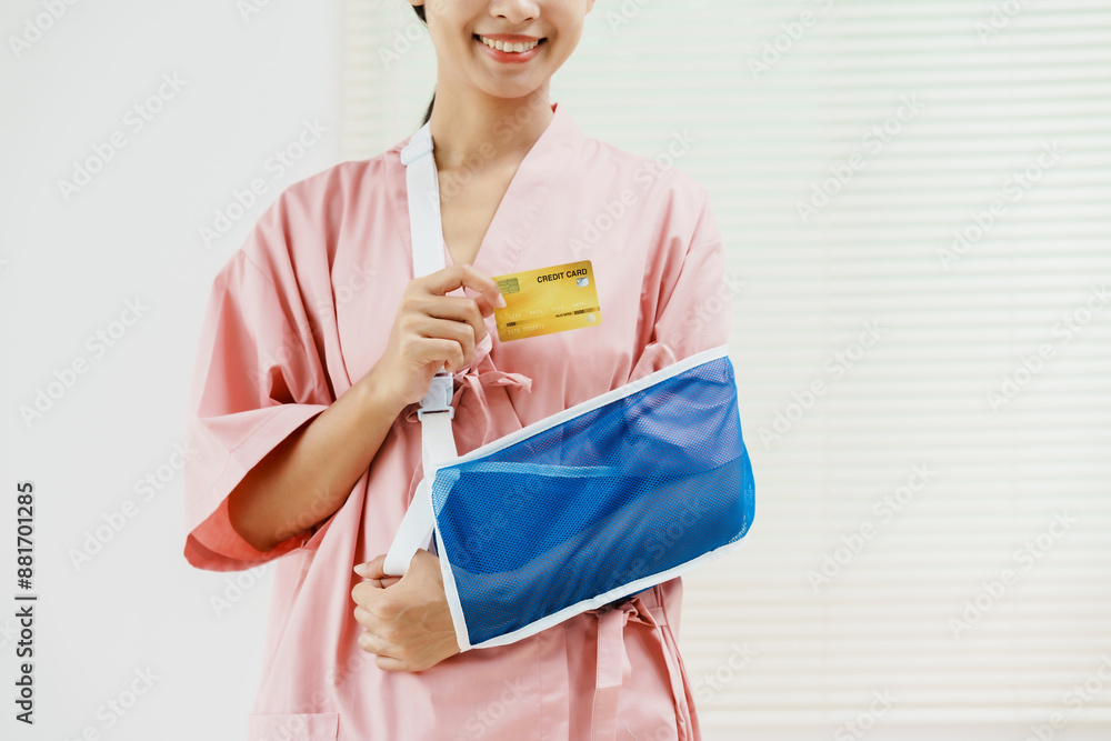 A young Asian female wearing a pink patient uniform stands isolated against a white wall background, showing confidence despite having her arm in a sling due to an injury. A young Asian female wearing a pink patient uniform stands isolated against a white wall background, showing confidence despite having her arm in a sling due to an injury.