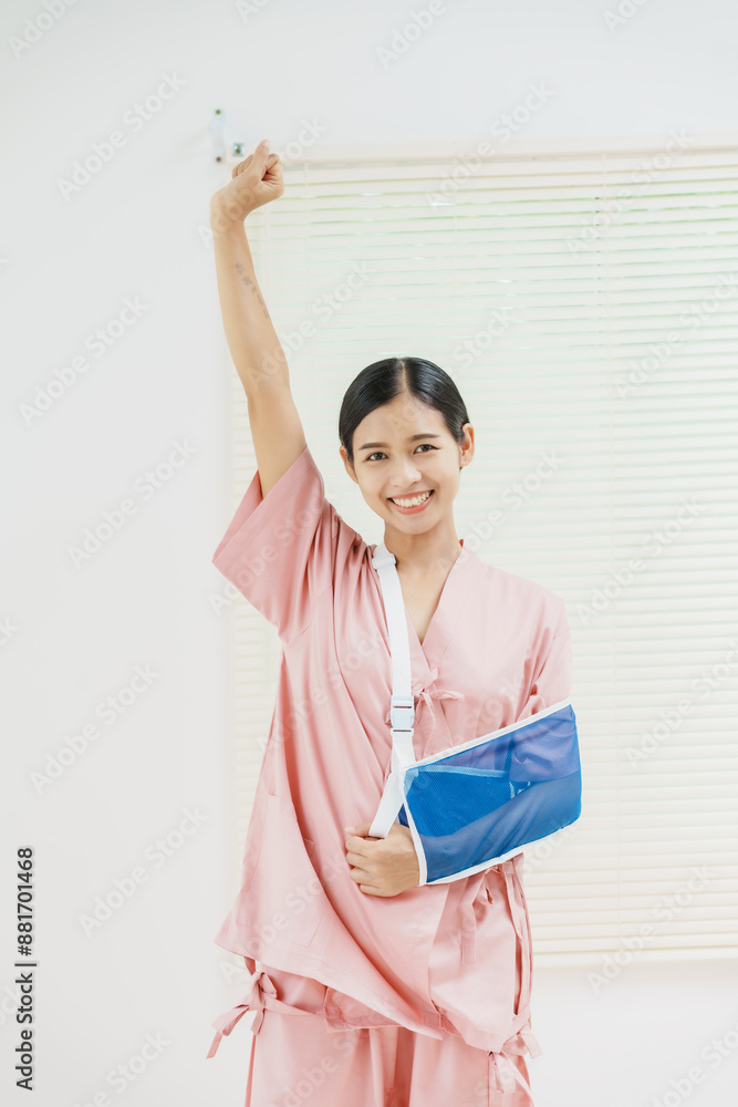 A young Asian female wearing a pink patient uniform stands isolated against a white wall background, showing confidence despite having her arm in a sling due to an injury. A young Asian female wearing a pink patient uniform stands isolated against a white wall background, showing confidence despite having her arm in a sling due to an injury.