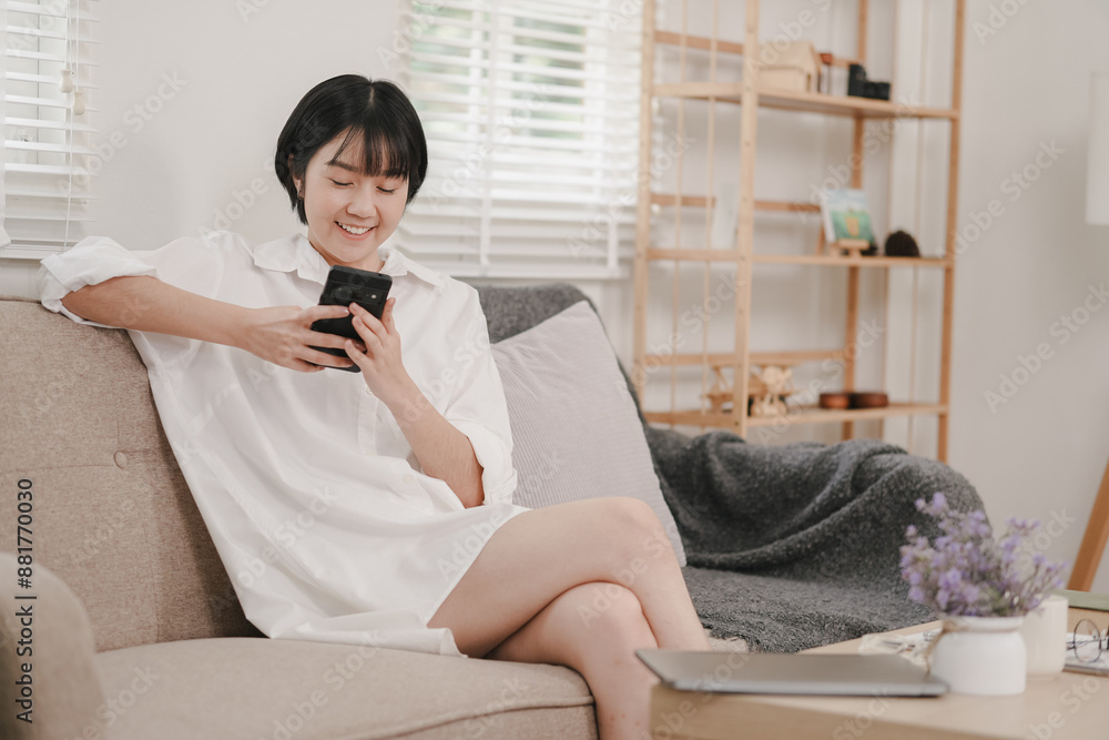 A woman is sitting on a couch and looking at her cell phone. She is smiling and she is enjoying herself. The room is decorated with various items, including a vase, a potted plant, and a laptop