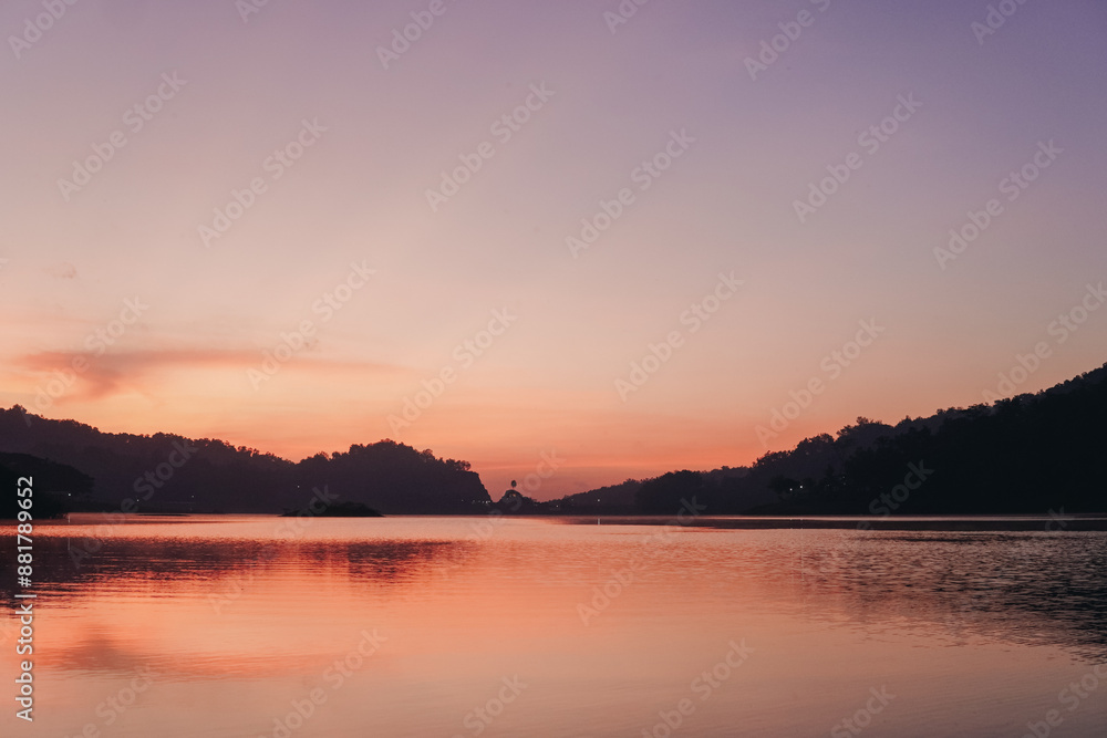 Fototapeta premium A beautiful sunset over Sermo lake, Yogyakarta, with a small island in the middle. The sky is a mix of pink and purple hues, creating a serene and peaceful atmosphere.