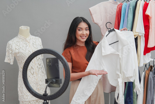 Attractive young Asian woman doing live stream online shopping pointing to the top white blouse she holds and presenting it to the camera.