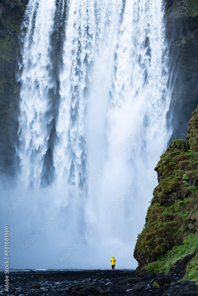 Stunning view of the Skógafoss Waterfall with a tourist wearing a yellow jacket. Skógafoss is one of the biggest waterfalls in Iceland, situated on the Skoga River in the south of Iceland.