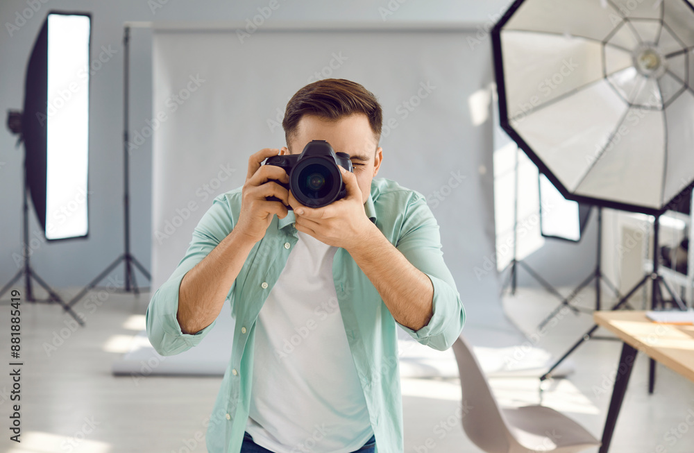 Professional photographer working in studio. Front view of young man in casual shirt and T shirt standing in studio with grey background and lighting equipment, holding camera, and taking photo Professional photographer working in studio. Front view of young man in casual shirt and T shirt standing in studio with grey background and lighting equipment, holding camera, and taking photo