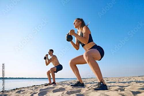 Fotografie Sportive young man and woman with sportive body, in black sportswear doing squats with kettlebell, intensive outdoor training on beach by the river