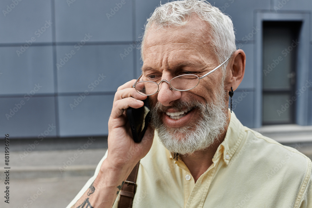 A mature, bearded man in glasses laughs as he talks on his phone outside a modern building. A mature, bearded man in glasses laughs as he talks on his phone outside a modern building.