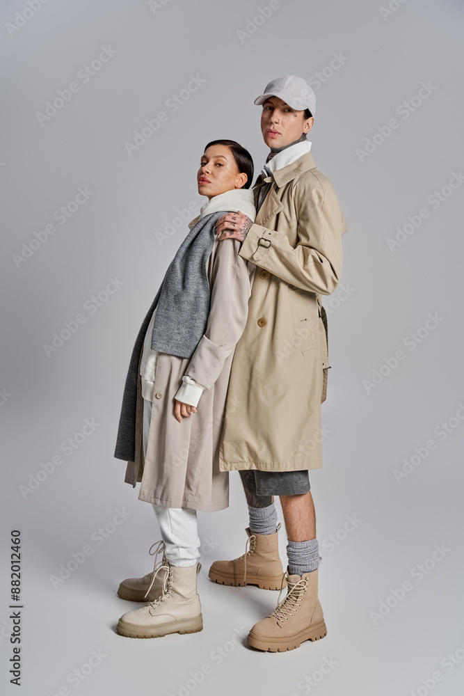 A young stylish couple stands side by side in trench coats, exuding sophistication and grace in a studio against a grey background. A young stylish couple stands side by side in trench coats, exuding sophistication and grace in a studio against a grey background.