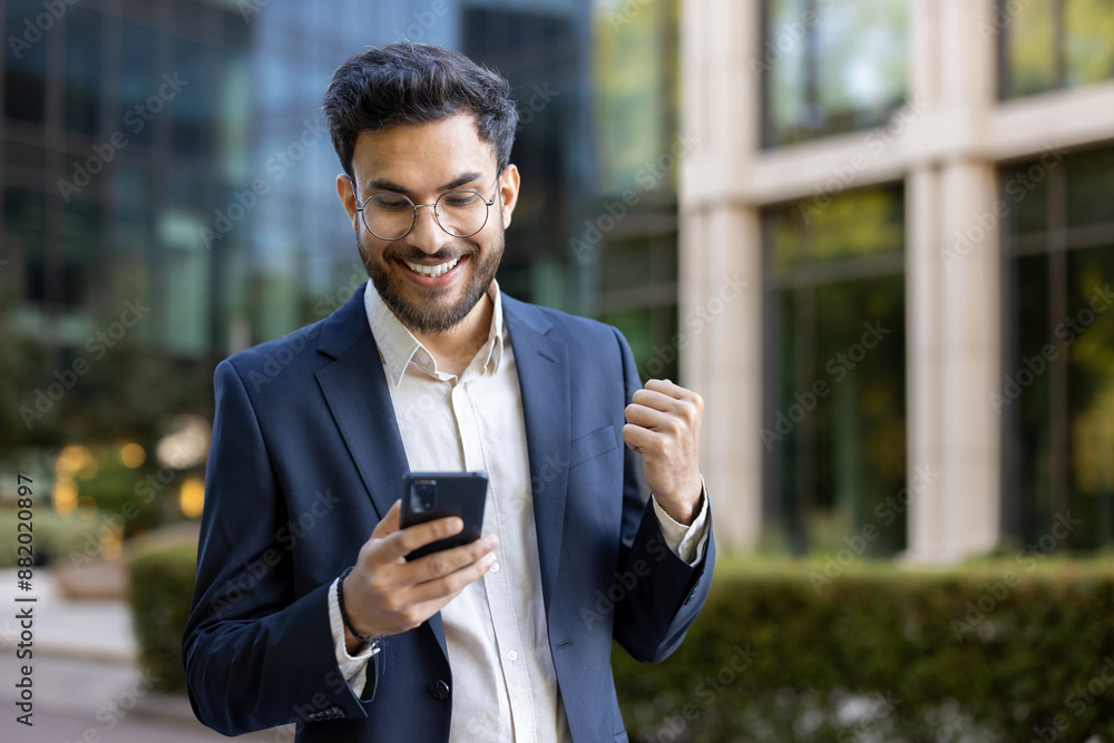 Young businessman in suit celebrating success while looking at smartphone outdoors. Urban background of modern office buildings. Focus on positive emotions, success, and modern technology. Young businessman in suit celebrating success while looking at smartphone outdoors. Urban background of modern office buildings. Focus on positive emotions, success, and modern technology.