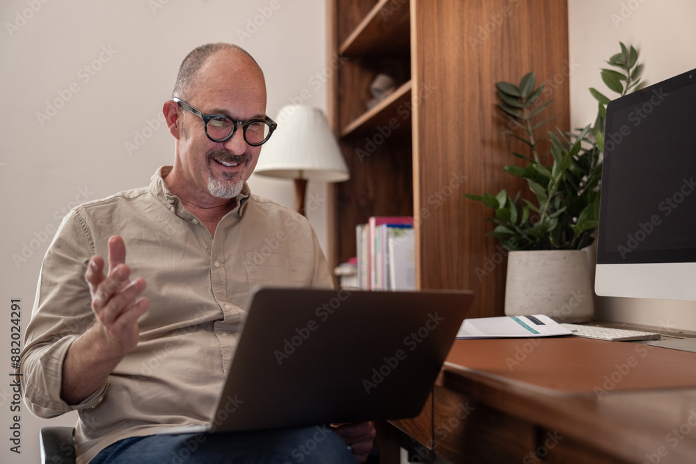 Mature man speaking on a videocall using a laptop in a home office Mature man speaking on a videocall using a laptop in a home office