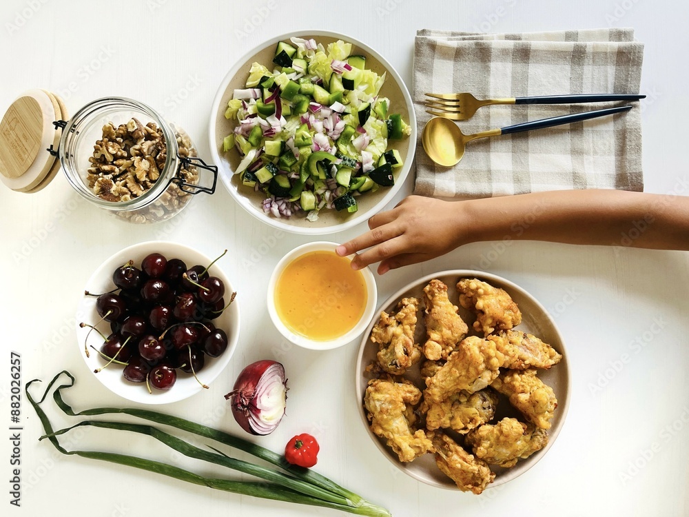 A child hand grab food. Dinner table with a plate of fried chicken, green salad, a bowl of cherry and cheese sauce on white wooden table A child hand grab food. Dinner table with a plate of fried chicken, green salad, a bowl of cherry and cheese sauce on white wooden table