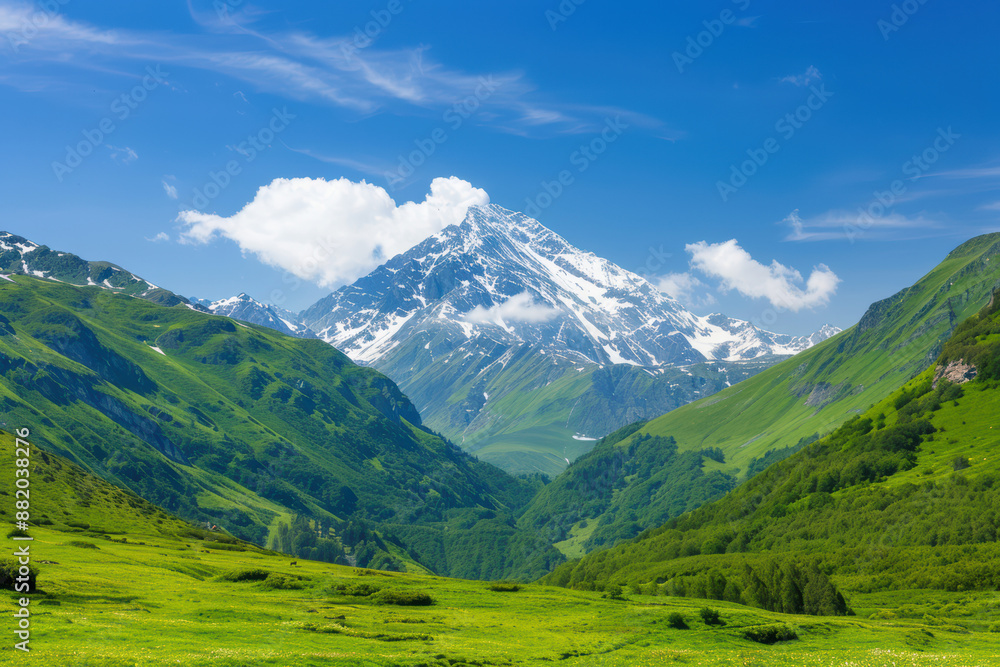 Obraz premium A majestic snow-capped mountain stands tall under a clear blue sky, surrounded by lush green hills and valleys. Perfect nature and travel scene.
