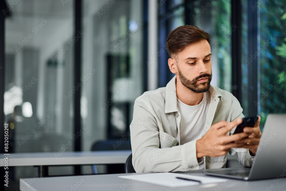 A focused man using a mobile phone while working at the office, over the laptop. A focused man using a mobile phone while working at the office, over the laptop.