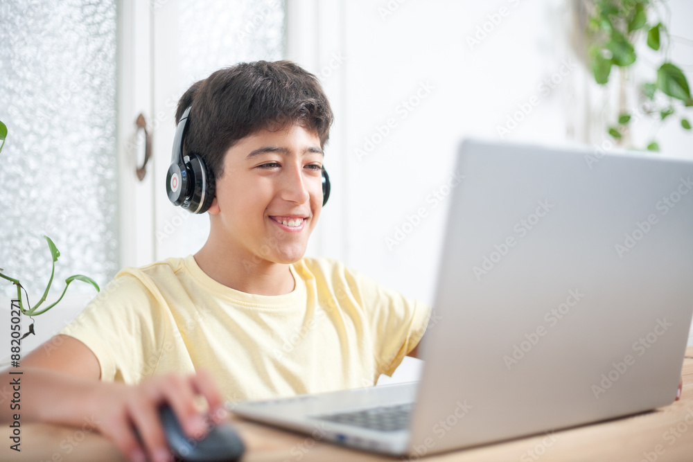 A cheerful young boy is sitting at a desk, wearing headphones and using a laptop computer in the living room of his home. A cheerful young boy is sitting at a desk, wearing headphones and using a laptop computer in the living room of his home.