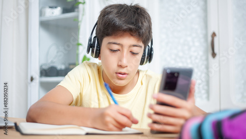 Boy with headphones writes on his notepad while checking his cell phone in his living room.