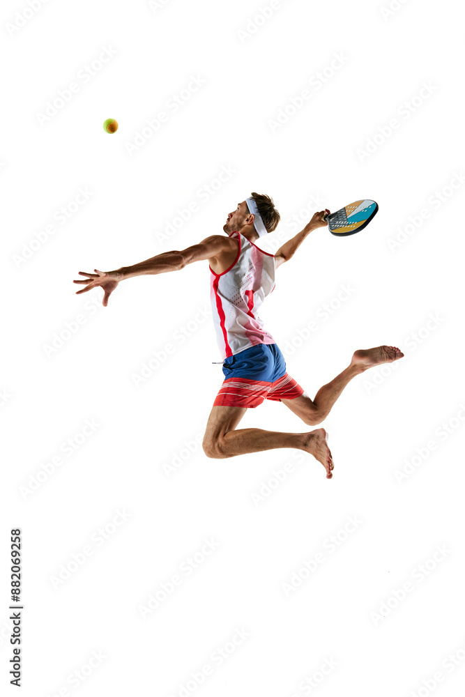 Concentrate man, beach tennis player in colorful sportswear hitting ball with racket in a jump isolated on white background. Concept of sport, active and healthy lifestyle, hobby, games, competition