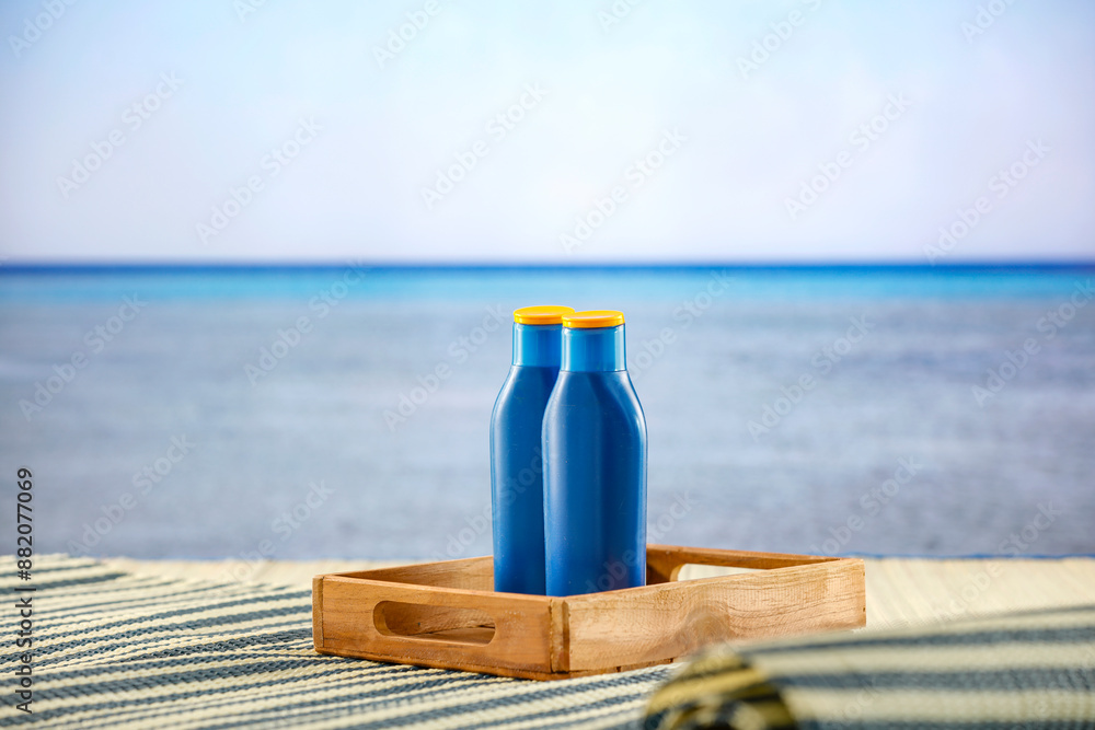 Sunscreen oils on a bech mat on a wooden deck with tropical summer sea and beach table background. Copy space for montage of products. Sunscreen oils on a bech mat on a wooden deck with tropical summer sea and beach table background. Copy space for montage of products.
