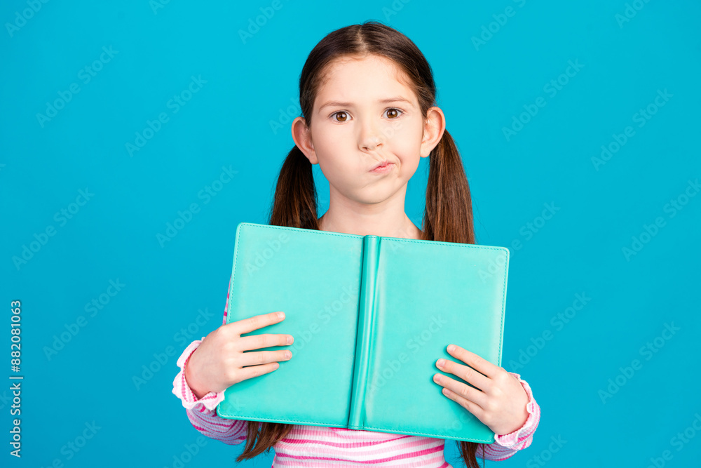 Photo of confused minded small girl with ponytails dressed striped shirt holding book make homework isolated on blue color background Photo of confused minded small girl with ponytails dressed striped shirt holding book make homework isolated on blue color background
