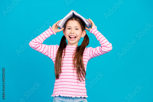 Photo of funny positive small girl with long ponytails dressed striped shirt holding book on head isolated on blue color background