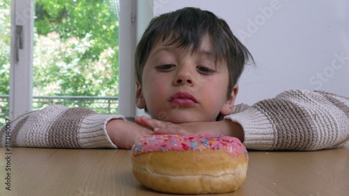 Schilderij op canvas A young boy peeking over a table, eyes fixed on a colorful sprinkled donut, conv