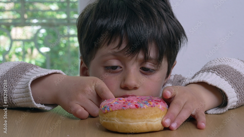 Boy pokes a pink frosted donut with colorful sprinkles on a wooden table, representing childhood desires and indulgence, curiosity and anticipation indoors Boy pokes a pink frosted donut with colorful sprinkles on a wooden table, representing childhood desires and indulgence, curiosity and anticipation indoors