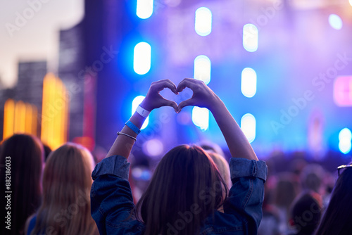 Back view of person forming heart shape with hands at music fest, surrounded by cheering crowd, vibrant stage lights blur in background, festive event atmosphere captures moment of joy at live concert