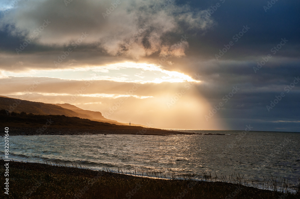 Dramatic coastal sunset with sun rays piercing through dark clouds, casting light over the ocean and rugged landscape Dramatic coastal sunset with sun rays piercing through dark clouds, casting light over the ocean and rugged landscape