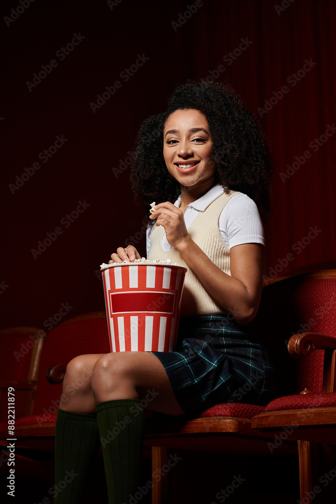 Naklejka premium A young woman enjoys a movie night, snacking on popcorn and reacting to the film with a big smile.