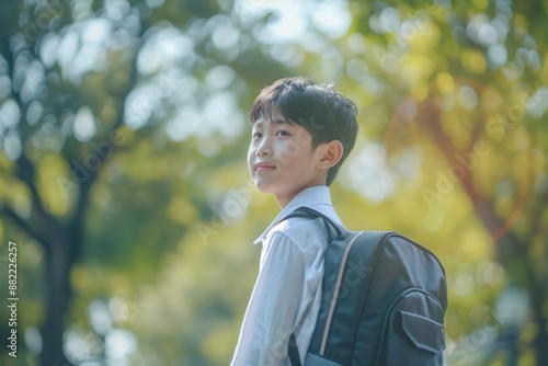 Asian teen boy wears classy school uniform in verdant park with backpack. Looks towards something unseen, ready for adventure. Green trees surround, leaves rustle gently.