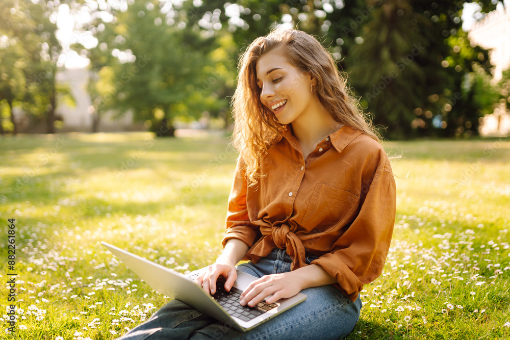 Young Woman sitting on grass ground, working on laptop pc computer in city park on green grass sunshine lawn outdoors. Mobile Office. Freelance business concept Young Woman sitting on grass ground, working on laptop pc computer in city park on green grass sunshine lawn outdoors. Mobile Office. Freelance business concept