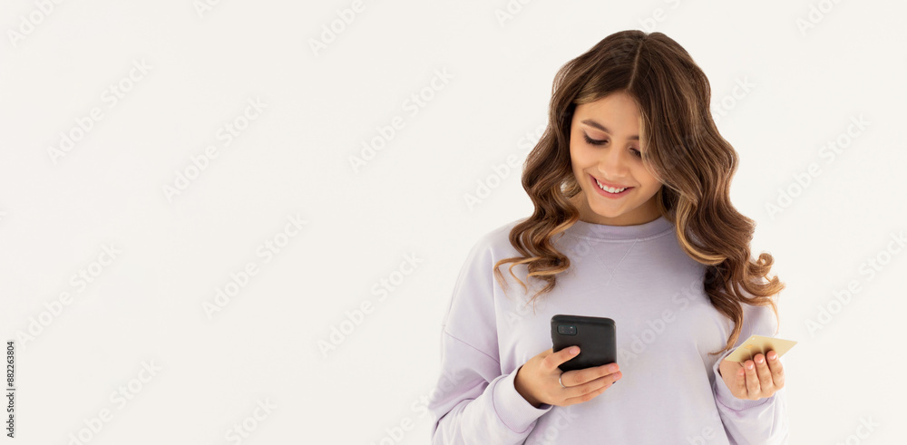 Cheerful young girl showing plastic credit card while using mobile phone isolated over white background, wearing purple sweatshirt Cheerful young girl showing plastic credit card while using mobile phone isolated over white background, wearing purple sweatshirt