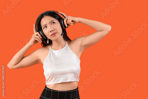 Thoughtful young beautiful Asian woman in headphones on orange background