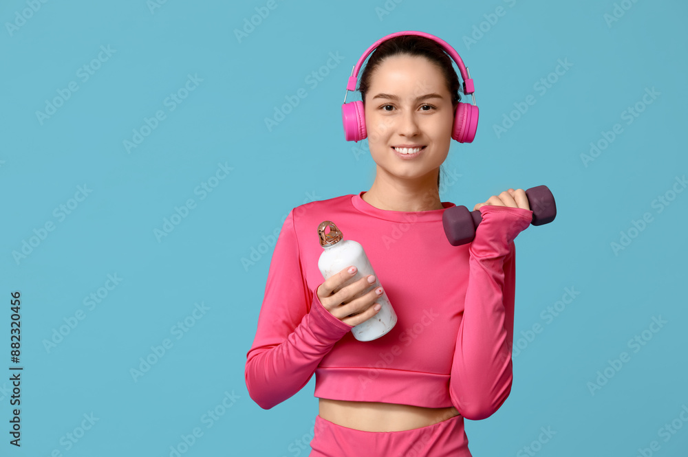 Young sporty woman in headphones with water bottle and dumbbell on blue background Young sporty woman in headphones with water bottle and dumbbell on blue background