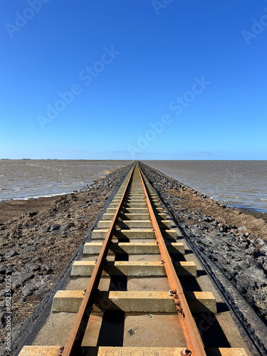 Straight railway tracks stretch along a stone dam or causeway into the sea toward the horizon under a clear blue sky.